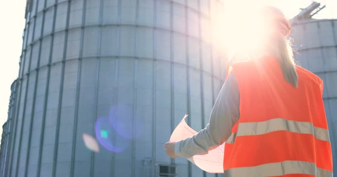 Rear of beautiful Caucasian female controller in uniform and helmet standing outdoor and holding graphs. Portrait of woman engineer in sunshine near big cisterns looking at plan. Factory concept