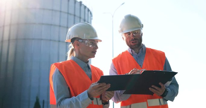 Portrait of Caucasian male and female constructors in helmets with tablet and planner speaking outdoors. Woman and man engineers in goggles discussing industry manufacturing plan. Factory concept