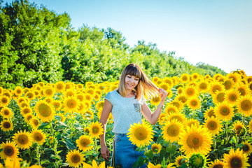 Fototapeta premium a young girl in a gray T-shirt and denim skirt stands in a bright yellow sunflower field. Happy girl smiling in a field of sunflowers