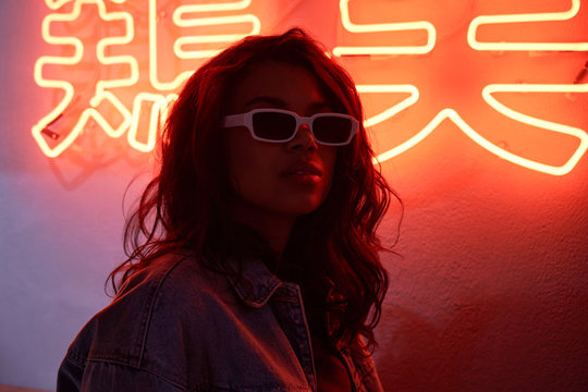Stylish cool fashion sexy African American young attractive hipster girl mixed race model wears trendy sunglasses stands in red neon backlight in night club. 80s 90s gen z retro style party nightclub