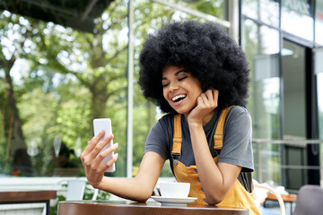 Happy African American young hipster gen z teen girl with Afro hair holding smart phone making video call, shooting social media blog talking with friend, watching video sitting at outdoor cafe table.