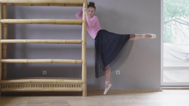 slender woman in pink sweater, black ballet skirt and pointe shoes doing exercises at home, adapting existing furniture for this