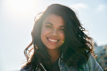 Smiling attractive African American teen generation z girl with dental smile looking at camera, outdoor headshot. Happy mixed race young woman posing on city street, face lit with sunshine, portrait.