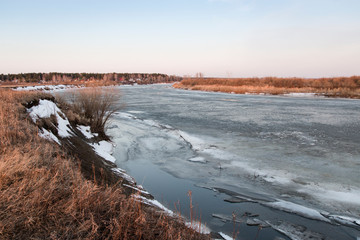 Landscape. Early spring morning. Gray ice on the river, the banks are covered with old yellow grass, blue sky. In the background there are houses and a forest.