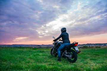 Obraz na płótnie Canvas Man sitting on a black motorcycle, wearing blue jeans, a black jacket and a black helmet, with his back turned against a landscape in the background.