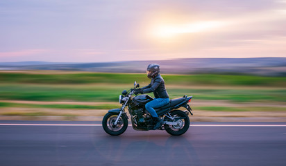 Plakat Man sitting on a black motorcycle, dressed in jeans and black jacket, moving on the road with the background out of focus.