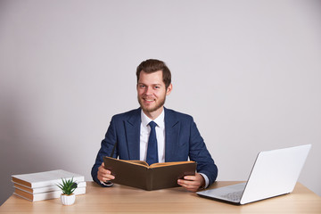 A businessman in a blue business suit sits at a wooden desk, holds a book, smiles and looks into the camera. White background.