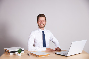 A businessman in a white shirt and blue tie sits at his desk and looks into the camera.