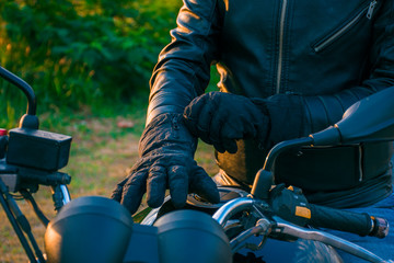 Obraz premium Man sitting on a black motorcycle, dressed in jeans and a black jacket, putting on black gloves with the landscape in the background.