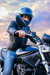 Fototapeta na wymiar Man sitting on a black motorcycle, wearing jeans, black jacket and black helmet with a background of the sky with clouds.