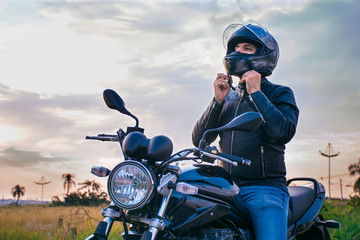 Man sitting on a motorcycle, wearing jeans and a black jacket, fastening his helmet with a landscape in the background. - obrazy, fototapety, plakaty