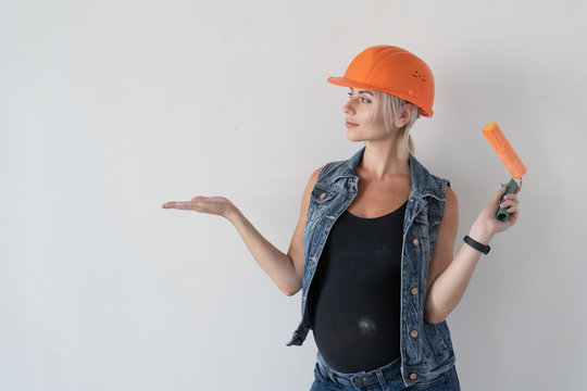 Young beautiful woman builder in an orange protective helmet on her head. Pregnant girl. holds a paint roller