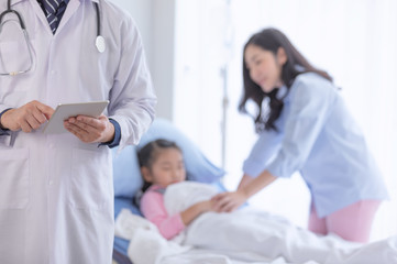 The doctor with tablet on hand for diagnosing the illness for a little girl on the hospital bed with mom beside.