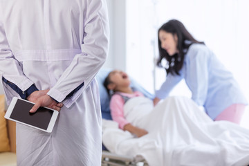 The doctor with tablet on hand for diagnosing the illness for a little girl on the hospital bed with mom beside.