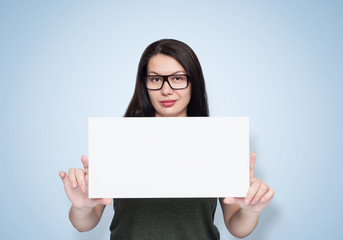 Naklejka premium Portrait of a cute smiling young girl in glasses holding a blank white banner in her hands, on light blue background. 