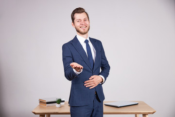 A businessman in a blue suit stands near a wooden office table and holds out an open hand to the camera, smiling. White background.