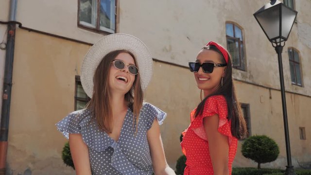 Two best cheerful tourists in a stylish look in sunglasses and summer dresses playfully dance and having fun in urban street,posing to camera. Blonde and brunette.