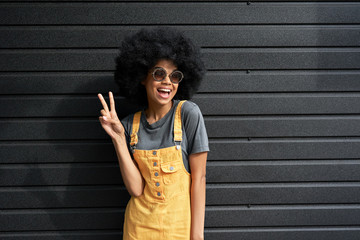 Fototapeta na wymiar Happy African American teen girl wearing stylish glasses, yellow trendy sundress with Afro hair showing peace hand sign on black background. Funky smiling fashion mixed race hipster woman portrait.