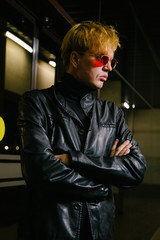 A stylish young man in a leather jacket stands with his arms crossed on the background of a corporate building.