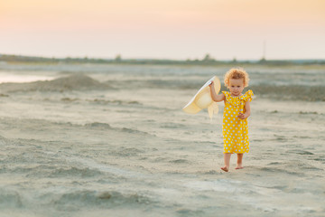 portrait of a little curly-haired blonde girl in a yellow dress with a flounce and a white hat on the background of the sandy desert at sunset in the evening