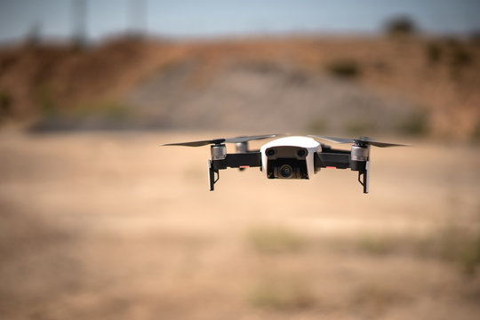 Drone blanco con cuatro h&eacute;lices volando a baja altura en un paisaje des&eacute;rtico, con su c&aacute;mara estabilizada apuntando hacia la c&aacute;mara
