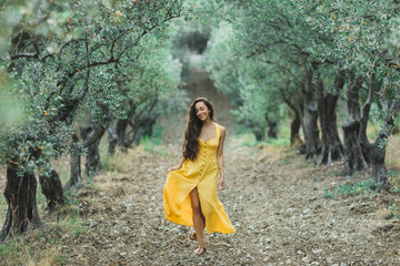 Young happy smiling woman walking in olive tree garden. Yellow linen summer dress. Trendy color and...