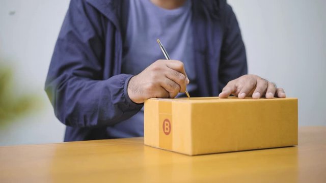 Fast post shipment service concept. young asian man customer writing address on parcel cardboard box at home office desk. 4K Resolution.