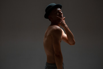 Stylish young guy actor in hat stands without shirt in studio isolated on black background