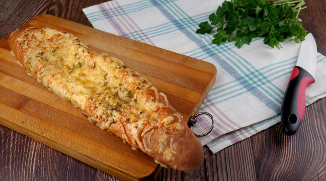 Homemade bread - baguette with cheese on top on a cutting board on a white towel next to a knife and green vegetables in the background.