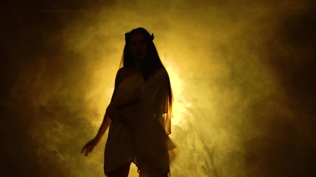Burning brunette is dancing in a nightclub in the dark and dynamic neon lights. Smoky background. Dancing go go. Silhouette girl in greek greece goddes dress and wreath high fashion.
