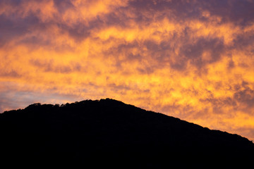 A fiery orange-red sunset. Ragged Cirrus clouds in the dark blue night sky. Black silhouette of a mountain. Amazing phenomenon. Background.