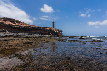 Rocky Atlantic coastline at low tide. In the background, the Punta Jandia lighthouse. Fuerteventura. Canary Islands. Spain.