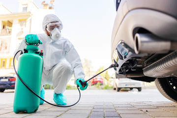 Man in white protective uniform with rubber gloves holding sprayer with disinfectant and spraying car. Protection from corona virus concept.