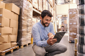 Young bearded smiling supervisor crouching in warehouse and using tablet to check on goods.