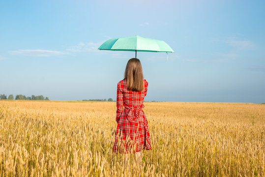 A young woman in a red dress and holding a green umbrella among the golden spikelets in a wheat field. Concept of protection, calmness and freedom