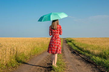 A young woman in a red dress and a green umbrella is walking along the road along a wheat field. Concept of protection, calmness and freedom