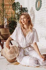 Summer fashion conception: fashionable woman wearing white crochet jumpsuit, many chains, holding straw hat, posing in stylish boho interior.  © Victoria Fox