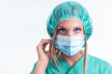 Nurse with blue eyes, green uniform, and mask, isolated in a white background, looks towards the camera, and fixes her hair inside the headgear.