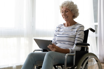 Happy old mature disabled woman sitting in invalid carriage, using computer tablet, enjoying...