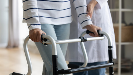 Close up middle aged woman holding hands on walking frame, trying making steps with physiotherapist assistance. Disabled retired old female patient doing exercises at rehabilitation procedure.