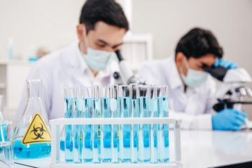 ..Focus on Solution Tubes in front of Two Male Scientists wear Face Mask working in Lab while Checking Solution by micro scope On Background...