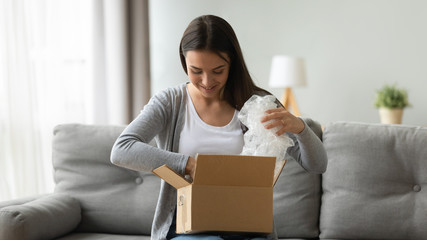 Happy young pretty woman sitting on comfortable sofa, unwrapping cardboard box with ordered online goods at home. Smiling millennial pleasant girl unpacking parcel with sale internet purchase.