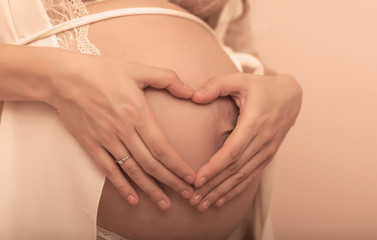 Closeup of pregnant woman holding her hands on her swollen belly shaping a heart, toned in pastel colors.