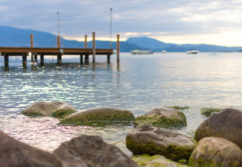 In the background a boardwalk leads into Lake Garda. In the foreground are large pebbles and stones overgrown with algae. The sky is cloudy.