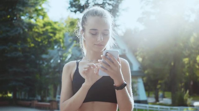 Close uo sporty girl using mobile phone stand at sunlight smile runner healthy workout fitness female outside sporty modern track mobile jogger athlete slow motion