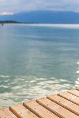 In the background, the mountains around Lake Garda are blurred. The focus is on the wooden boards of a footbridge in the foreground. The sky is cloudy.