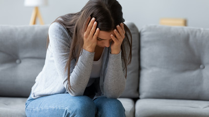 Frustrated young woman holding head in hands, feeling depressed alone at home. Unhappy doubtful hopeless millennial girl lost in negative thoughts, having headache migraine, inner emptiness.