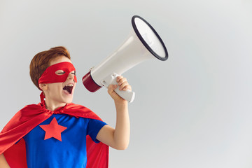 Cute boy dressed as superhero shouting into megaphone on grey background. Child in superman costume...