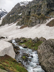 Fototapeta na wymiar water stream flowing in between a huge rock on one side and a snow glacier on other side in an epic Indian Himalayan Valley.