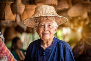 Smiling elderly Thai female street vendor wearing local hats and Mauhom shirt of local handicraft souvenir shops selling tourists in Thailand.